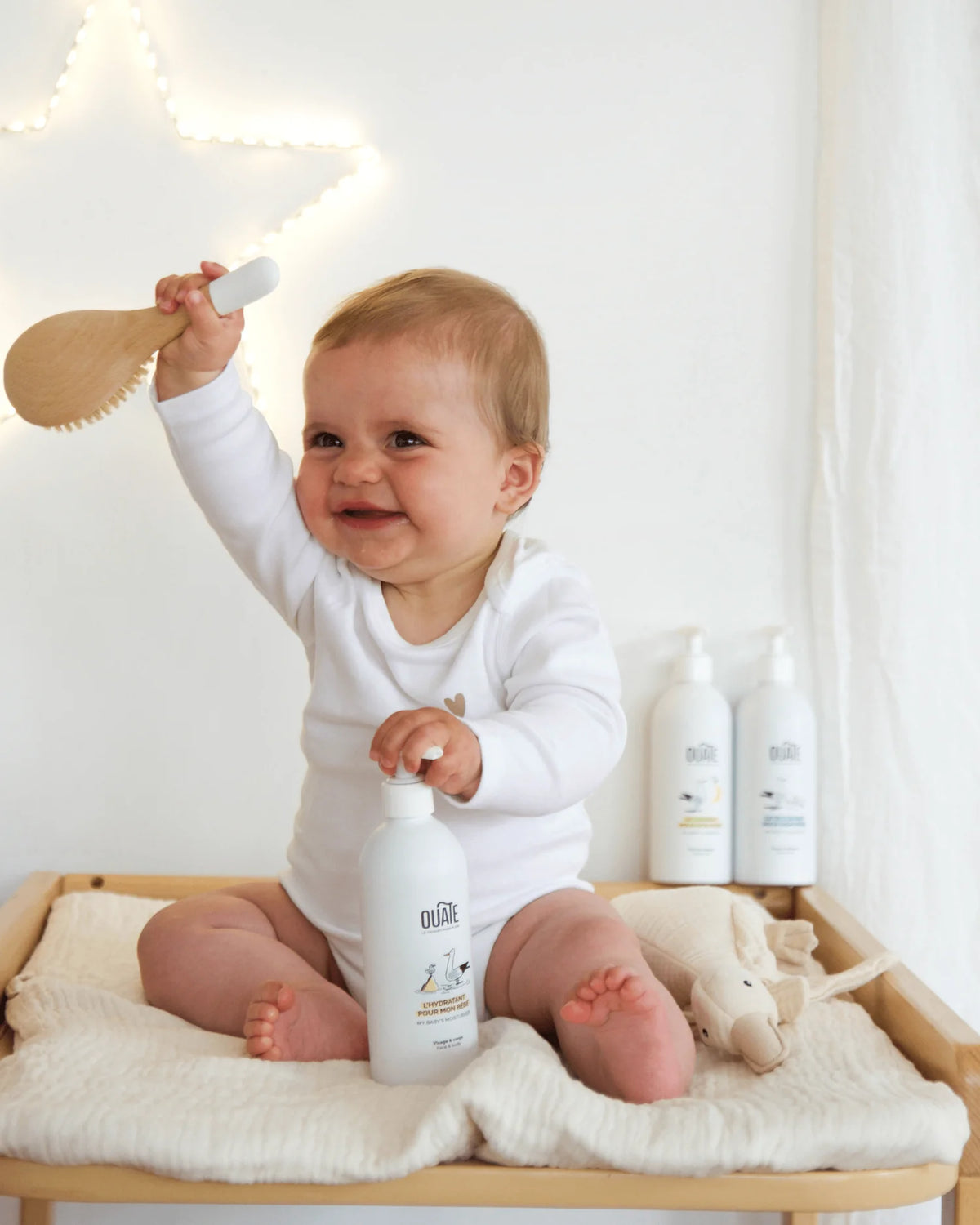 Bébé rieur assis sur une table à langer, brosse à cheveux en main, flacons Ouate pour bébé à côté
